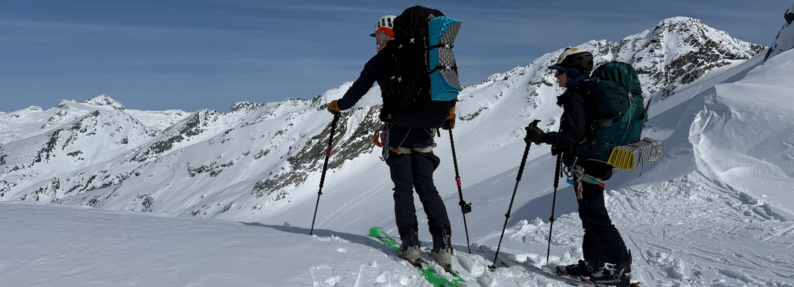 Two skiers carrying heavy packs across a glaciated alpine panorama